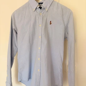 Blå slim fit Ralph Lauren skjorta size S - Mycket bra skick. Snygg ljusblå skjorta från Ralph Lauren i slim fit-modell. Skjortan har klassisk krage, långa ärmar och knappar framtil. Tillverkad i 100% bomull för skön känsla och bra passform.