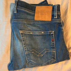 Replay Hyperflex Grover jeans blå - Snygga Replay Hyperflex Grover jeans i klassisk blå tvätt med raka ben och tydliga slitningar. Jeansen har fem fickor, kontrastsömmar och en läderpatch bak i midjan. Materialet är stretchigt för extra komfort och passformen är straight fit. Passar för dig som är 173-175