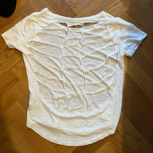 Vit t-shirt med skeleton design på ryggen - använde den till kent !Vit t-shirt i bomull med korta ärmar och rund halsringning. Baksidan har ett coolt slitsat mönster som ger en edgy och unik look. Perfekt för dig som vill sticka ut med en kreativ och trendig stil.