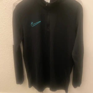 Svart Nike Dri-Fit half zip tröja - Svart långärmad Nike Dri-Fit tröja med halv dragkedja och turkos broderad swoosh på bröstet. Tröjan har vita paneler på axlar och ärmar, perfekt för träning eller chill. Tillverkad i funktionsmaterial som andas och transporterar bort fukt. Från JD