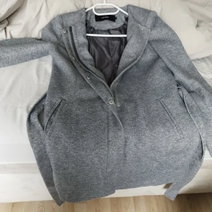 Grå kappa från Vero Moda med dragkedja - Snygg grå kappa från Vero Moda med stilren design, dragkedja och knappar framtill. Jackan har två fickor på sidorna och är fodrad på insidan för extra komfort. Perfekt för dig som gillar en klassisk look med modern touch.