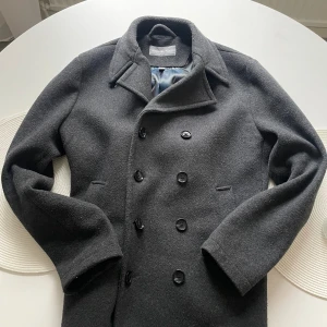 Mörkgrå kavaj från Michael Kors - Stilren mörkgrå skepparkavaj från Michael Kors i stretchig ullblandning. Jackan har dubbelknäppning med svarta knappar, klassisk krage och två fickor framtill. Perfekt för dig som vill ha en tidlös och snygg look under höst och vinter. S men passar M