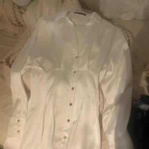 Vit skjortklänning i satin från Zara - Snygg vit skjortklänning från Aveny i glansigt satinmaterial. Klänningen har långa ärmar, guldiga knappar framtill och på manschetterna samt markerad midja för en stilren look. Perfekt för dig som gillar klassiska och trendiga plagg.