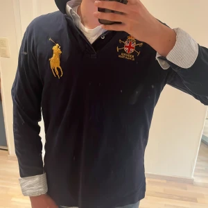 Mörkblå pikétröja från Ralph Lauren - Säljer en mörkblå långärmad pikétröja från Ralph Lauren med stora broderade detaljer, bland annat en gul polospelare på bröstet och brittisk flagga med texten 'Mercer RL Polo Team'. Stor gul siffra 3 på ryggen och ärmen. Klassisk krage och knappar vid halsen. Svinfet tröja och en bra färg som man kan använda till vardags men även finare tillställningar! Ny pris 2295