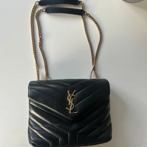 Svart axelväska från Saint Laurent - Säljer en svart axelväska från Saint Laurent med klassiskt YSL-emblem i guld framtill. Väskan är quiltad i skinn och har en kedjerem med läderdetaljer. Perfekt storlek för det viktigaste och levereras med originalbox och dustbag.