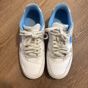 Nike Air Force 1 Low vit/blå - Säljer ett par Nike Air Force 1 Low i vitt skinn med ljusblå detaljer och swoosh. Skorna har klassisk snörning, rund tå och platt sula. Insidan är fodrad i blått tyg för extra komfort. Perfekta sneakers för dig som gillar en clean och sportig look.