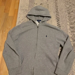 Grå hoodie från Polo Ralph Lauren XL - Klassisk grå hoodie från Polo Ralph Lauren i storlek XL. Tröjan har dragkedja, huva och två fickor framtill. Broderad marinblå logga på bröstet. Mjuk och skön bomullsblandning, perfekt för chill dagar.
