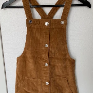 Brun manchesterklänning FB Sister XS - Supersnygg brun kortklänning i manchester från FB Sister, storlek XS. Klänningen har knappar framtill, två fickor och coola korsade axelband. Perfekt för dig som gillar retrovibbar och vill sticka ut med en unik look.