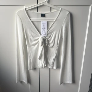 Vit långärmad topp med knyt från Gina Tricot - Säljer en stilren vit långärmad topp från Gina Tricot i storlek XS. Toppen har v-ringning och snygg knytning framtill som ger en trendig look. Materialet är mjukt och stretchigt, perfekt för att skapa en clean och fräsch outfit.