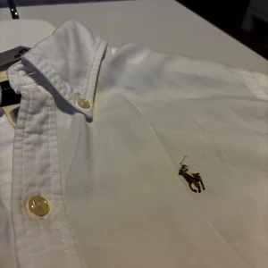 Vit skjorta från Ralph Lauren - Vit skjorta från Ralph Lauren med klassisk button-down krage och broderad logga på bröstet. Stilren och passar till många olika tillfällen. Slim fit och lite liten i storleken. Passar M/S.