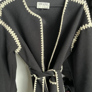 Svart kappa med broderade detaljer AWARE - Svart kappa från AWARE i storlek S med snygga ljusa broderade kanter och knytskärp i midjan. Jackan har två fickor framtill och raka, långa ärmar. Perfekt för dig som gillar stilrena plagg med unika detaljer.