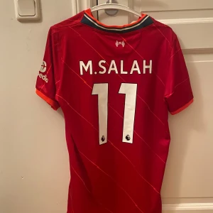 Liverpool Salah matchtröja Nike L - Liverpool FC hemmatröja med M. Salah och nummer 11 på ryggen. Röd med tunna diagonala ränder, vita och orange detaljer, Premier League-märke på ärmen och sponsorloggor. Tillverkad i Dri-FIT-material som andas. Perfekt för dig som älskar fotboll och vill sticka ut.