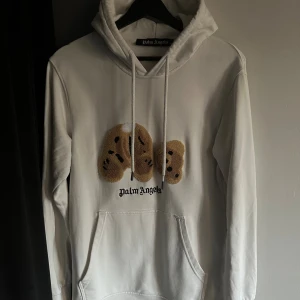 Vit Palm Angels hoodie  - Säljer en vit hoodie från Palm Angels med ett broderat nallemotiv på bröstet och svart Palm Angels-tryck under. Tröjan har huva med snörning och en stor magficka. Perfekt för dig som gillar streetwear och coola detaljer.