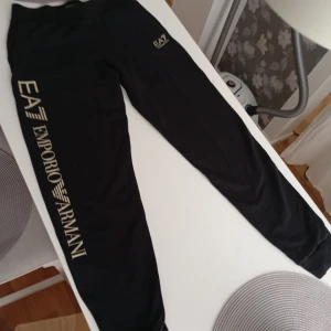 Svarta EA7 Emporio Armani mjukisbyxor - Svarta mjukisbyxor från EA7 Emporio Armani med stort guldfärgad logotryck längs ena benet och mindre logga på höften. Byxorna har resår i midjan och är tillverkade i mjukt material, perfekta för en chill och sportig look.