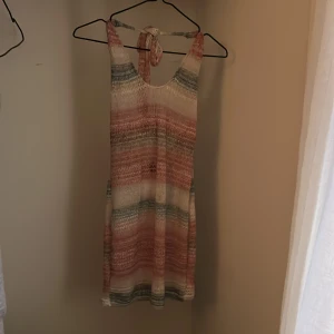 Randig stickad sommarklänning - Superfin stickad kortklänning med breda ränder i beige, rosa och grönt. Klänningen har knyt i nacken och är ärmlös, perfekt för varma dagar. Den har en avslappnad passform och känns både trendig och bekväm.