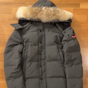 Canada Goose Wyndham - Säljer en grå vinterjacka från Canada Goose med ikonisk röd och vit patch på ärmen. Jackan har en fluffig päls på huvan, knappar framtill och en unik design. Perfekt för kalla vinterdagar och har en riktigt snygg, klassisk look.