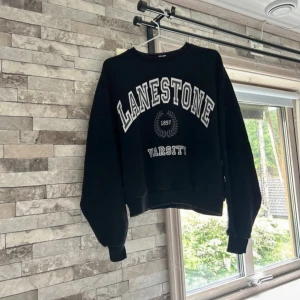 Svart sweatshirt Lanestone Varsity - Cool svart sweatshirt från Nelly  med trycket 'LANESTONE VARSITY 1897' i vitt framtill. Tröjan har en loose passform, ribbade muddar och rund hals. Perfekt för en avslappnad och sportig stil.