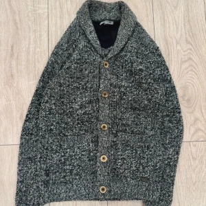 Cardigan från Poolman - Säljer en gråmelerad stickad kofta från Poolman med snygg sjalkrage och träknappar. Koftan har två fickor framtill och är perfekt för kyliga dagar. Den är stickad i grovt garn.