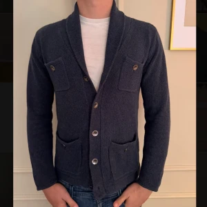 Mörkblå kofta från GANT med fickor - Snygg mörkblå kofta från GANT i 100% bomull. Koftan har fyra fickor med knappar framtill och en bred krage som ger en chill och klassisk vibe. Perfekt att slänga över en t-shirt för en avslappnad look. Knäppning med fyra knappar framtill.