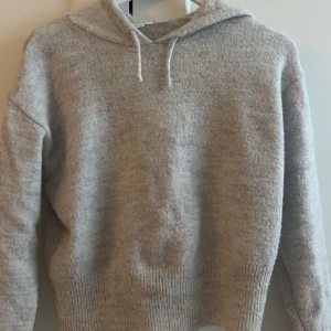 Grå stickad hoodie med huva - Mysig grå stickad hoodie med huva och dragsnören. Tröjan har ribbade muddar vid ärmslut och nederkant, perfekt för kyliga dagar. Enkel och stilren design som passar till det mesta. Använd ett par gånger men bra skick!