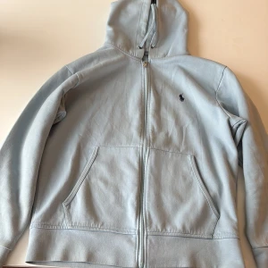 Ljusblå hoodie från Polo Ralph Lauren - Ljusblå hoodie från Polo Ralph Lauren med dragkedja framtill, två stora fickor och klassisk broderad logga på bröstet. Tröjan har huva med snörning och är tillverkad i mjuk bomull. Perfekt för chill dagar eller när du vill ha en clean look.