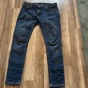 Mörkblå jeans från Nudie Jeans, 33/30 - Säljer ett par mörkblå jeans från Nudie Jeans i klassisk femficksmodell. Jeansen har orangea sömmar, raka ben och snygga detaljer på bakfickorna. Tillverkade i slitstarkt bomullstyg och passar perfekt till en avslappnad stil.