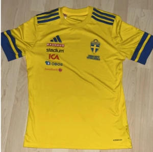 Svenska landslagets fotbollströja Adidas - Gul fotbollströja från Adidas med blå detaljer och tryck från Landslagets Fotbollsskola. Tröjan har korta ärmar med blå ränder, flera sponsorloggor på bröstet och klassiskt landslagsmärke. Tillverkad i lätt och ventilerande funktionsmaterial. GÅVA FRÅN SVERIGES FOTBOLLFÖRBUND DÅ DEN ÄR ÄKTA