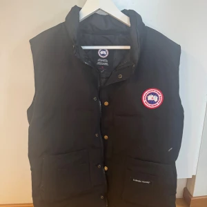 Svart dunväst från Canada Goose - Svart dunväst från Canada Goose med klassisk logga på bröstet, hög krage och stora fickor framtill. Västen har både dragkedja och tryckknappar. Perfekt för lager-på-lager och riktigt snygg till streetwear-stilen.