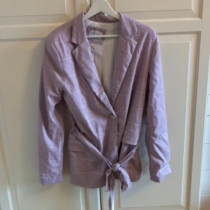 Ljuslila kavaj med knyt från Bershka - Säljer en ljuslila kavaj från Bershka med diskret rutigt mönster. Kavajen har en knapp framtill, knytband i midjan och två fickor. Materialet känns mjukt och lätt. Snygg passform och trendig färg! Storlek M. I fint skick. 