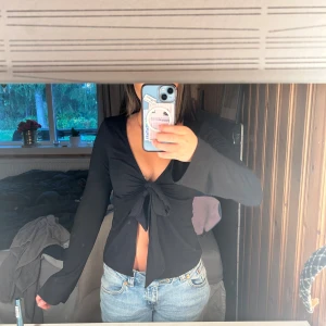 Svart omlottopp med knyt från Gina Tricot - Säljer en svart omlottopp från Gina Tricot i storlek S. Toppen har långa ärmar, djup v-ringning och snygg knytning framtill som ger en croppad look. Perfekt att styla med jeans eller kjol för en trendig vibe.