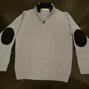 Ljusblå halvzip-tröja med armbågslappar - Snygg ljusblå stickad tröja från Barneys med svart krage och halv dragkedja. Tröjan har svarta armbågslappar som ger en cool touch. Perfekt för dig som gillar klassisk men trendig stil.