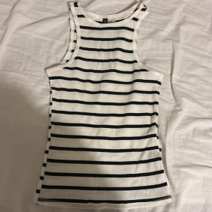 Randig ribbad linnetopp i vitt och svart - Säljer en fräsch ribbad linnetopp från H&M med svarta horisontella ränder på vit botten. Toppen är ärmlös med rund hals och har en slim passform som sitter snyggt mot kroppen. Perfekt för dig som gillar enkel och stilren design.