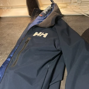 Mörkblå vindjacka från Helly Hansen - Säljer en mörkblå vindjacka från Helly Hansen med vit HH-logga på bröstet. Jackan har dragkedja framtill, hög krage med mjukt foder och en bröstficka med dragkedja. Perfekt för blåsiga dagar och outdoor-äventyr.