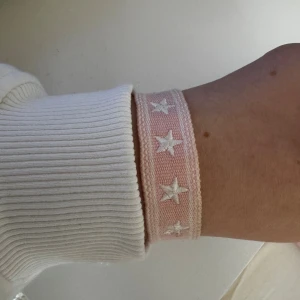 Rosa armband med stjärnor och tofsar - Säljer ett brett, vävt armband i ljusrosa med vita broderade stjärnor. Armbandet har justerbar knytning med två tofsar i ändarna för en extra detalj. Perfekt accessoar för att ge din outfit en söt och trendig touch.💘                                                            Tror den är köpt i Cypern men minns inte riktig, sparsamt använt.                                                           Skickar ditt köp inom 24h!💘