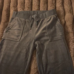 Gråa mjukisbyxor Juicy Couture S - Blåa mjukisbyxor från Juicy Couture i mjuk sammet med bred resår i midjan och snörning med metalländar. Stora fickor fram och bak med knappdetaljer. Perfekta för en chill och avslappnad look.