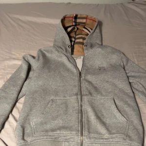 Grå zip-hoodie från Burberry M - Grå hoodie från Burberry med dragkedja och klassiskt rutigt mönster i huvan. Broderad logga på bröstet och två stora fickor framtill. Perfekt för vintern.