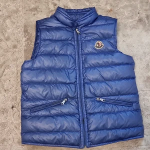 Blå dunväst från Moncler - Snygg blå dunväst från Moncler med quiltad design och två snedställda fickor med dragkedja. Västen har hög krage och Moncler-logga på bröstet. Perfekt att slänga över en hoodie för extra värme och stil.