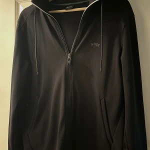 Svart hoodie med dragkedja från BOSS - Svart hoodie från BOSS med hel dragkedja, huva med snörning och diskret BOSS-logga på bröstet. Tröjan har långa ärmar och två fickor framtill. Tillverkad i mjuk bomull för en skön och avslappnad stil. Köpt för 1200kr, säljer för 1000kr. Den är som ny!