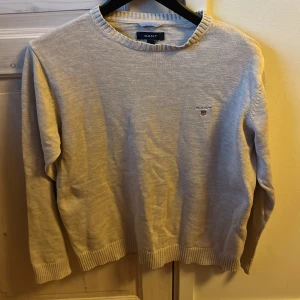 Beige stickad tröja från GANT - Säljer en stilren beige stickad tröja från GANT i storlek M. Tröjan har rund halsringning, ribbade muddar och diskret GANT-logga broderad på bröstet. Perfekt för lager-på-lager och enkel att matcha med jeans eller chinos.