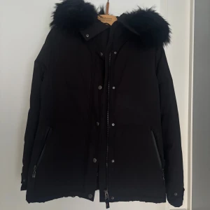 Svart dunjacka med pälskrage från Hollies - Snygg svart dunjacka från Hollies med stor fluffig pälskrage på huvan. Jackan har dragkedja och knappar framtill, två fickor med dragkedja och en klassisk badge på ärmen. Perfekt för kalla dagar och riktigt stilren look.