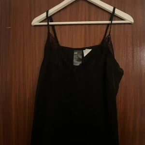 Svart linnetopp med spets från H&M - Svart linnetopp från H&M med tunna axelband och snygga spetsdetaljer vid halsringningen och längs sidorna. Toppen är ärmlös och har en lätt, flowy passform. Materialet är polyester med spets i polyamid och elastan. Perfekt för dig som gillar stilrena och trendiga plagg.