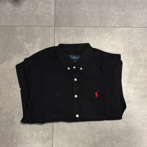 Svart skjorta från Ralph Lauren - Svart skjorta från Ralph Lauren med klassisk krage och vita knappar framtill. På bröstet finns den ikoniska röda Polo-loggan broderad. Skjortan har lång ärm och är gjord i mjuk bomull, perfekt för en clean och stilren look. Passar mer som S än M enligt mig.