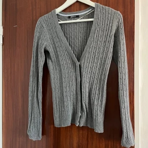 Grå ribbad kofta från Gina Tricot - Grå kofta från Gina Tricot i storlek S. Den är ribbstickad med smala vertikala ränder och har v-ringning samt knäppning framtill. Långärmad och i mjukt stickat material, perfekt att slänga över en topp när det är lite kyligt.