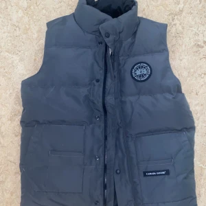 Mörkgrå dunväst från Canada Goose - Snygg mörkgrå dunväst från Canada Goose med hög krage, två stora fickor framtill och diskret logga på bröstet. Västen har dragkedja och tryckknappar samt klassisk quiltad design. Perfekt för lager-på-lager under kyliga dagar.