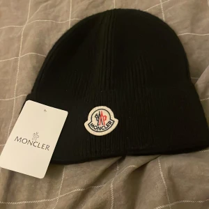 Svart Moncler mössa i ull - Snygg svart mössa från Moncler med klassisk logotyp framtill. Mössan är ribbstickad och tillverkad i mjuk ull, perfekt för kyliga dagar. Enkel och stilren design som passar till allt.