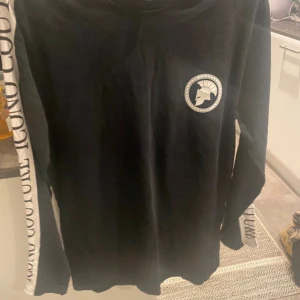 Svart långärmad tröja från Iconic Couture - Svart långärmad tröja från Iconic Couture med vit logga på bröstet och vita paneler längs ärmarna med svart text. Tröjan har en clean och stilren look, perfekt för dig som gillar streetwear och tydliga detaljer. Materialet känns som mjuk bomull.