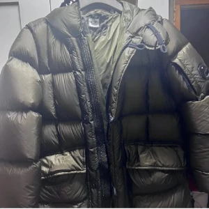 Olivgrön dunjacka från C.P. Company - Säljer en olivgrön dunjacka från C.P. Company med fet pufferlook, huva och klassisk linsdetalj på ärmen. Jackan har stora fickor, dragkedja och knappar framtill. Perfekt för kalla dagar när du vill hålla stilen och värmen. 