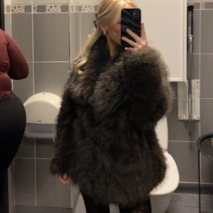 Mörkgrå fluffig pälsjacka - Säljer en superfluffig och mörkgrå pälsjacka med bred krage. Jackan har en riktigt lyxig känsla och är perfekt för dig som vill sticka ut och hålla dig varm under kalla dagar. Passformen är perfekt och är enkel att styla till olika outfits. Är inte äkta!
