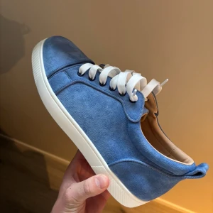 Blå mockasneakers från Louboutin - Snygga blå sneakers i mocka från Louboutin med klassisk röd sula och vita snören. Skorna har rund tå, platt vit sula och diskreta detaljer som ger en clean look. Perfekta för dig som vill sticka ut med stilrena och lyxiga sneakers.