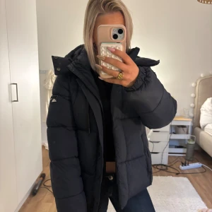Mörkblå pufferjacka från Superdry - Säljer en svart pufferjacka från Superdry med huva och dragkedja. Jackan är vadderad och har en klassisk pufferlook med breda kanaler. Märkeslogga på ärmen och insidan. Perfekt för kalla dagar och riktigt snygg till jeans. Köpte för 2000kr
